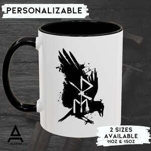 May include: White ceramic mug with a black rim and handle. The mug features a black and white illustration of a raven with a stylized symbol on its chest. The text "2 SIZES AVAILABLE 11OZ & 15OZ" is printed on the bottom of the mug.