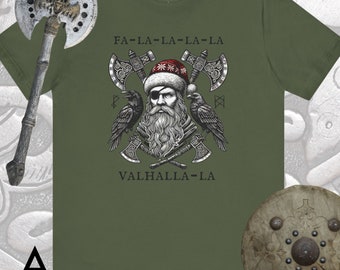 Può includere: T-shirt verde oliva con una grafica di un uomo barbuto con cappello da Babbo Natale e benda sull'occhio, con asce incrociate e corvi. Il testo "FA-LA-LA-LA-LA" e "VALHALLA-LA" sono sopra e sotto la grafica. Un'ascia e uno scudo sono sullo sfondo.
