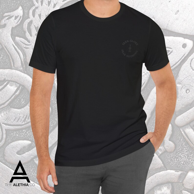 May include: Black crew neck t-shirt with the text "SEEK GLORY FEAR NOTHING" in a circular design on the upper left chest. The shirt is a solid black color and is made of a soft material. The Alethia Co. logo is visible in the bottom left corner.