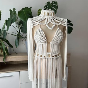 Op de afbeelding: Een witte gehaakte jurk met een franjesrok en een bijpassend topje met een kralenhalsband. De jurk is op een mannequin en heeft een gouden kralenriem.