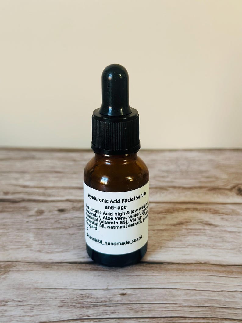 Op de afbeelding: Een klein amberkleurig glazen flesje met een zwarte druppelaar. Het flesje is gelabeld met "Hyaluronic Acid Facial Serum anti-age" en vermeldt ingredi&euml;nten zoals hyaluronzuur, alo&euml; vera, water, vitamine B5, ylang-ylang etherische olie, haverextract en conserveermiddelen. Het etiket bevat ook de tekst "Vandiotti handmade soaps".