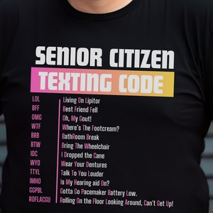 Op de afbeelding: Zwart T-shirt met witte tekst die "Senior Citizen Texting Code" luidt en een lijst met veelvoorkomende sms-afkortingen voor senioren en hun betekenis.