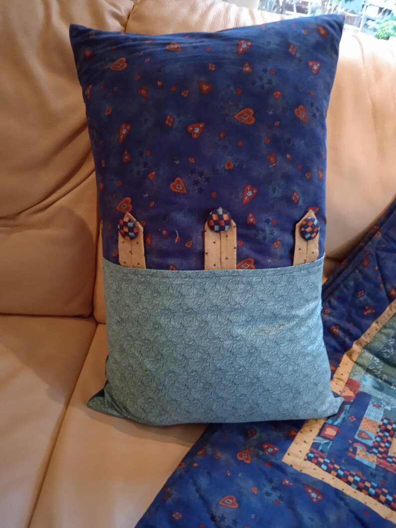 May include: A blue and green pillow with a heart pattern and three buttons. The pillow has a pocket on the back.