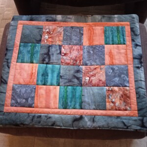 Pode incluir: Uma colcha de retalhos com um esquema de cores cinza, azul-esverdeado e laranja. A colcha &eacute; feita de quadrados de diferentes tecidos e &eacute; bordada com um tecido cinza escuro.