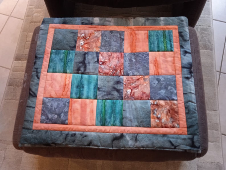 Pode incluir: Uma colcha de retalhos com um esquema de cores cinza, azul-esverdeado e laranja. A colcha &eacute; feita de quadrados de diferentes tecidos e &eacute; bordada com um tecido cinza escuro.