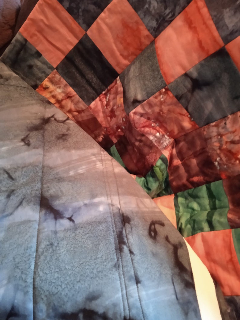 Pode incluir: Uma colcha com um design de patchwork que apresenta quadrados de diferentes cores e padr&otilde;es. A colcha &eacute; feita de tecido com efeito tie-dye. As cores incluem tons de marrom, laranja, verde e azul.