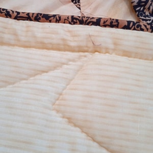 May include: Close-up of a cream-colored quilted fabric with thin horizontal stripes. The fabric has a decorative stitch pattern and a brown and black patterned border.