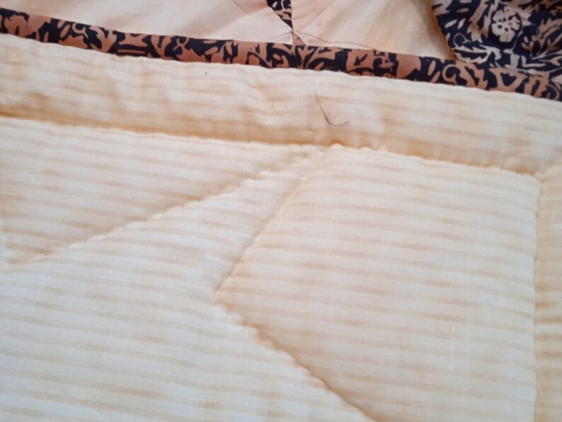 May include: Close-up of a cream-colored quilted fabric with thin horizontal stripes. The fabric has a decorative stitch pattern and a brown and black patterned border.