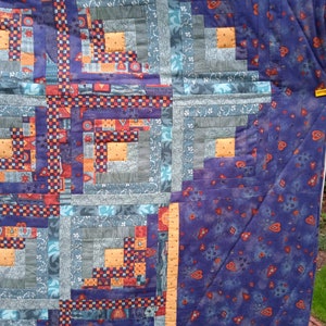 May include: A quilt with a log cabin pattern in shades of blue, orange, red, and purple. The quilt is made of various fabrics with different patterns and textures. The quilt is hanging on a clothesline.
