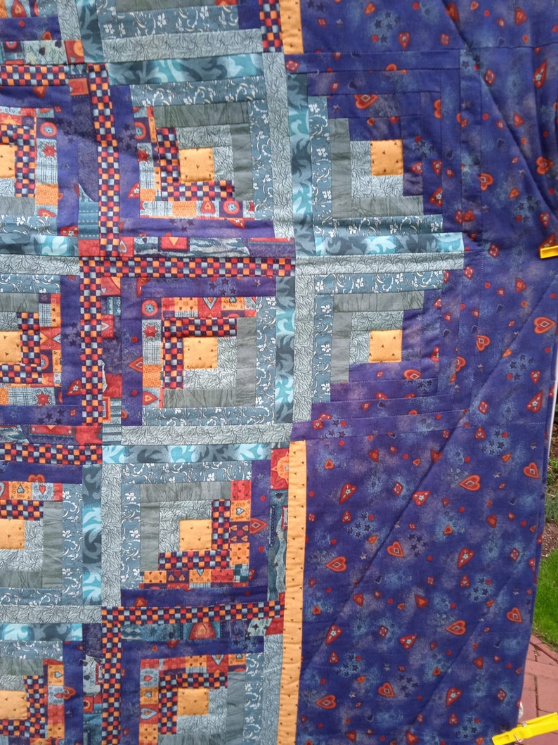 May include: A quilt with a log cabin pattern in shades of blue, orange, red, and purple. The quilt is made of various fabrics with different patterns and textures. The quilt is hanging on a clothesline.