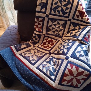May include: A blue and red patchwork quilt with a white background. The quilt is made up of squares with different patterns, including stars, pinwheels, and diamonds. The quilt is folded and draped over a brown armchair.