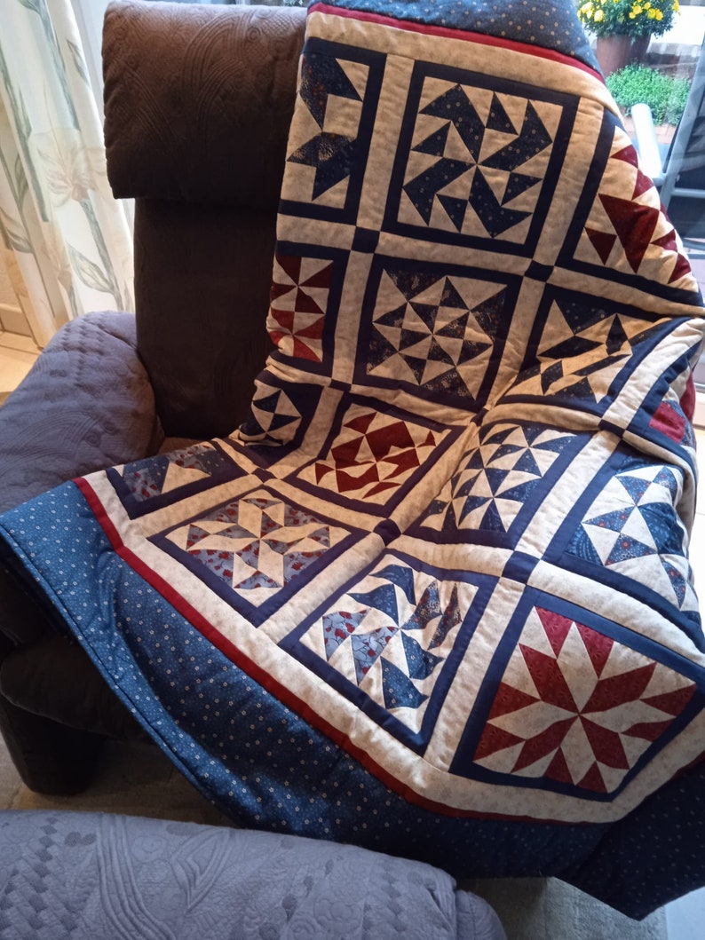May include: A blue and red patchwork quilt with a white background. The quilt is made up of squares with different patterns, including stars, pinwheels, and diamonds. The quilt is folded and draped over a brown armchair.