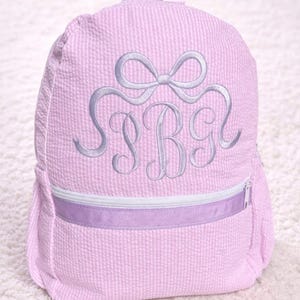 Pode incluir: Mochila rosa em tecido seersucker com laço e bordado de monograma em lilás claro. A mochila tem fecho de correr e uma pega superior. O monograma inclui as letras SBG.