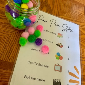 Pode incluir: Um cartão branco com o texto "Pom Pom Store" e uma lista de atividades com números correspondentes. As atividades incluem "Bedtime story", "Candy Treat", "Get a toy", "One TV Episode" e "Pick the movie". Cada atividade é ilustrada com uma pequena imagem. Um frasco de vidro transparente cheio de pompons coloridos está sobre uma superfície de madeira.