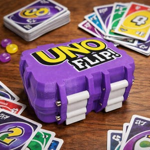 May include: A purple UNO FLIP! card game box with white latches and the game's logo in yellow and white. Several UNO cards are scattered around the box on a wooden surface, along with a stack of cards and some purple and yellow tokens.