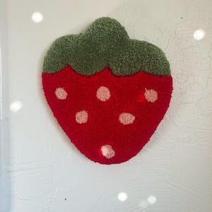 May include: A handmade strawberry-shaped rug with a red body and green stem. The rug features light pink seed details and a textured, tufted design. This decorative rug is suitable for a child's room or as a whimsical accent piece.