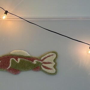 May include: A pink and green fish-shaped rug hangs on a white wall with a string of lights above it.