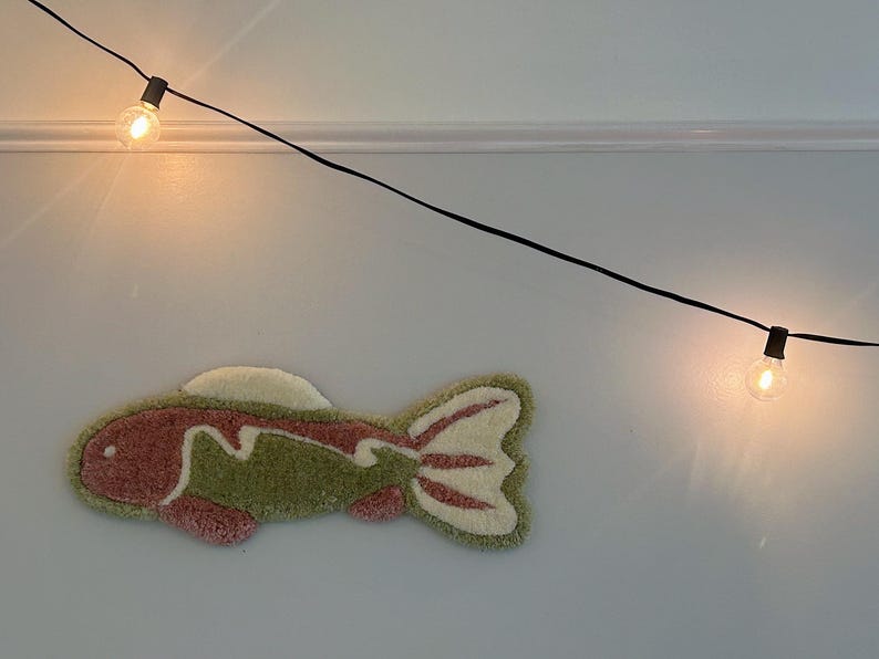 May include: A pink and green fish-shaped rug hangs on a white wall with a string of lights above it.