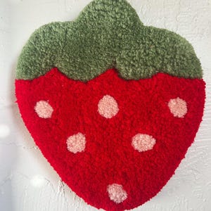 May include: A red strawberry-shaped rug with a green leafy top and pink seed details. The rug is made of a textured, plush material, and is designed to resemble a strawberry fruit.