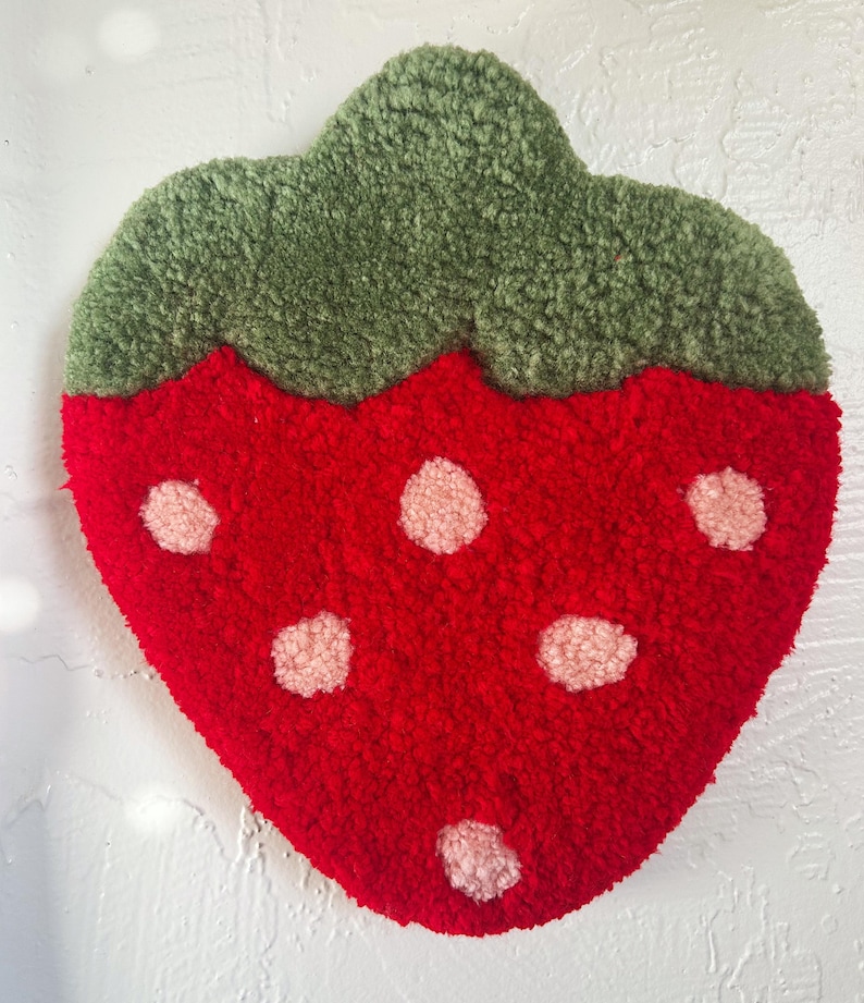 May include: A red strawberry-shaped rug with a green leafy top and pink seed details. The rug is made of a textured, plush material, and is designed to resemble a strawberry fruit.