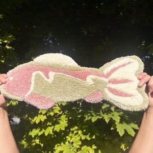 May include: A pink and green fish-shaped rug made of fluffy yarn. The rug is held up in front of a green leafy background.