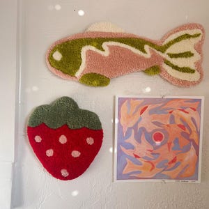 May include: A wall display featuring a pink and green fish-shaped rug, a red strawberry-shaped rug with white dots, and a square abstract painting with orange, red, and blue hues. The items are mounted on a white wall.