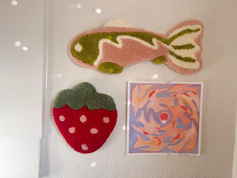 May include: A wall display featuring a pink and green fish-shaped rug, a red strawberry-shaped rug with white dots, and a square abstract painting with orange, red, and blue hues. The items are mounted on a white wall.