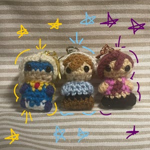 May include: Three handmade crochet doll keychains. The dolls have different hair colors and outfits, including blue, brown, and maroon. Each doll has a keychain ring attached to the top. The background has drawn stars and lines.