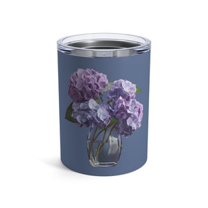 Op de afbeelding: Een roestvrijstalen tumbler met een blauwe achtergrond en een bloemenontwerp. Het ontwerp toont een boeket van paarse en blauwe hortensia's in een glazen vaas.