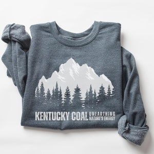 May include: Dark heather gray sweatshirt with a white mountain range and forest graphic. The text "Kentucky Coal Unearthing Nature's Energy" is printed below the graphic.