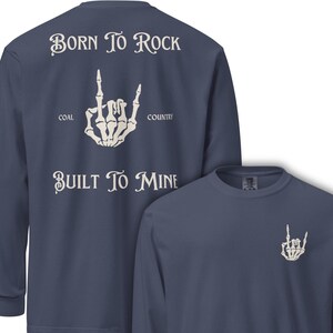 May include: A navy blue long-sleeve shirt with a white skeleton hand graphic making a rock and roll hand gesture. The shirt has the text "BORN TO ROCK" and "BUILT TO MINE". The shirt also has the text "COAL COUNTRY".