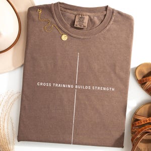 May include: A brown t-shirt with the text "CROSS TRAINING BUILDS STRENGTH" in white. A vertical white line bisects the text. A gold necklace with a pendant rests on the shirt. The shirt is next to a hat and sandals.