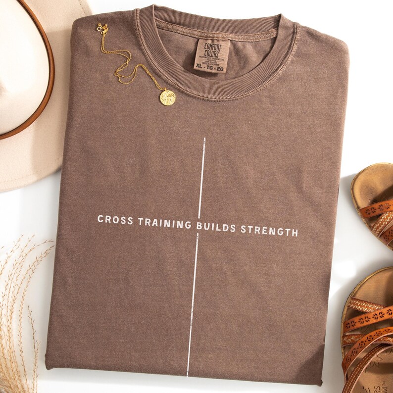 May include: A brown t-shirt with the text "CROSS TRAINING BUILDS STRENGTH" in white. A vertical white line bisects the text. A gold necklace with a pendant rests on the shirt. The shirt is next to a hat and sandals.