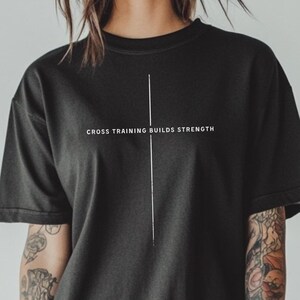 May include: Black t-shirt with the text "CROSS TRAINING BUILDS STRENGTH" in white. A vertical white line is centered on the shirt. The shirt has short sleeves and a crew neck.