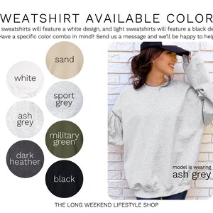 May include: A light grey sweatshirt is shown, with a model wearing the ash grey color. The image displays a color chart with options including white, sand, sport grey, military green, dark heather, and black. Text reads "SWEATSHIRT AVAILABLE COLORS".