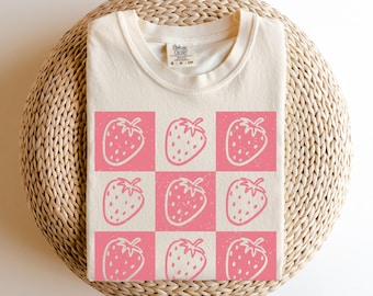 May include: Ivory-colored t-shirt featuring a grid pattern of pink strawberries. The strawberries have white outlines and are set against a pink and white checkered background. The shirt is made of a soft material and has a crew neck.