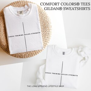 May include: Two white t-shirts and a sweatshirt are displayed. Each garment has a black vertical line and the text "CROSS TRAINING BUILDS STRENGTH." The top t-shirt is on a woven surface. The text "COMFORT COLORS TEES GILDAN SWEATSHIRTS" is at the top.