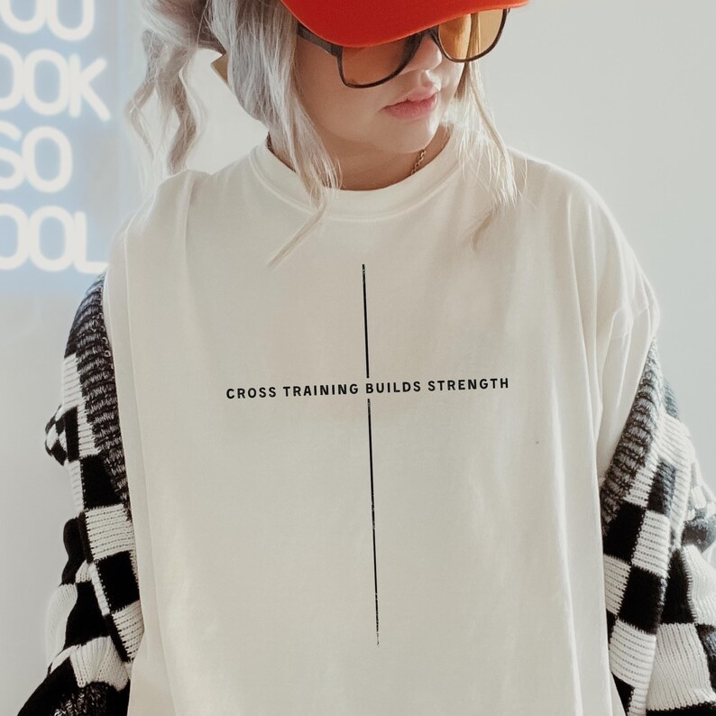 May include: A white t-shirt with the text "CROSS TRAINING BUILDS STRENGTH" in black. A vertical black line is centered on the shirt. The person is wearing an orange cap and a black and white checkered sweater.