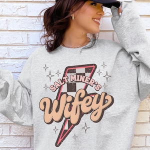 May include: A light gray sweatshirt with the text "Salt Miner's Wifey" in a retro font, with a lightning bolt graphic. The lightning bolt has a checkered pattern and pink accents. Four small star shapes surround the text.