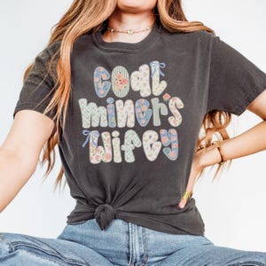 May include: Dark gray t-shirt with the text "Coal Miner's Wifey" in a patchwork design. The letters are decorated with floral and gingham patterns in pastel colors, with small blue bows.