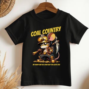 May include: Black t-shirt featuring a cartoon mouse miner with a pickaxe and a lamp. The text "COAL COUNTRY" is above the mouse, and "MY DADDY AND HIS CREW KEEP THE LIGHTS ON!" is below.