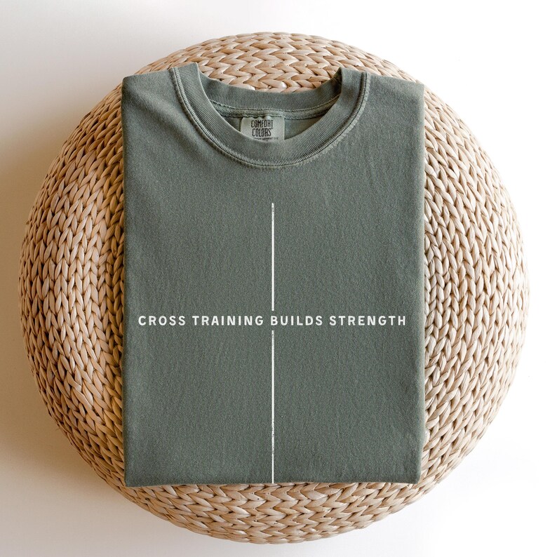 May include: A sage green t-shirt with the text "CROSS TRAINING BUILDS STRENGTH" in white. The shirt is folded and placed on a woven, circular surface. The shirt has a crew neck and short sleeves.