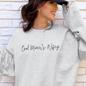 May include: A light gray sweatshirt with the text "Coal Miner's Wifey" in black script. The sweatshirt has long sleeves and a crew neck. The image shows the sweatshirt on a person.