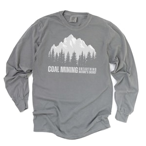 May include: A gray long-sleeve shirt featuring a white mountain and forest graphic. The text "COAL MINING UNEARTHING NATURE'S ENERGY" is printed below the graphic. The shirt is made of a soft material.
