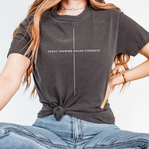 May include: Dark gray t-shirt with the text "CROSS TRAINING BUILDS STRENGTH" in white. A vertical white line is centered on the shirt. The shirt is knotted at the waist.