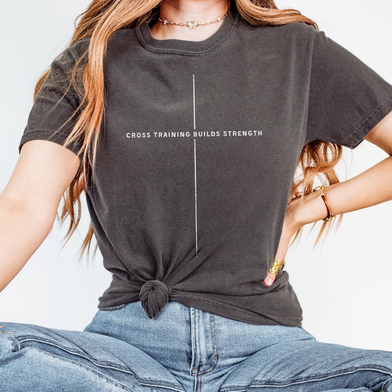 May include: Dark gray t-shirt with the text "CROSS TRAINING BUILDS STRENGTH" in white. A vertical white line is centered on the shirt. The shirt is knotted at the waist.