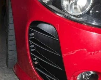 Puede incluir: Primer plano de la parte delantera de un coche rojo, que muestra un faro, una rejilla de ventilación negra y un neumático. La pintura del coche muestra signos de desgaste. La imagen destaca el diseño y los detalles del vehículo.