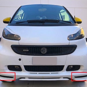Puede incluir: Smart blanco con parrilla negra y espejos laterales amarillos. La parte delantera del coche presenta una parrilla negra con el logotipo de Brabus y un gran emblema B. El coche tiene un parachoques delantero deportivo con rejillas de ventilación.