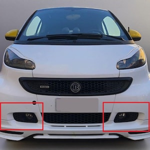 Peut inclure: Une Smart blanche avec une calandre noire et des feux antibrouillard. La voiture a des rétroviseurs latéraux jaunes et un pare-chocs avant noir. Le devant de la voiture porte le logo Brabus.