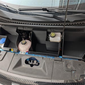 May include: A car engine bay with open hood, showing black plastic storage compartments and various engine components. Includes a coolant reservoir and a brake fluid reservoir. The engine bay is visible with its internal parts.